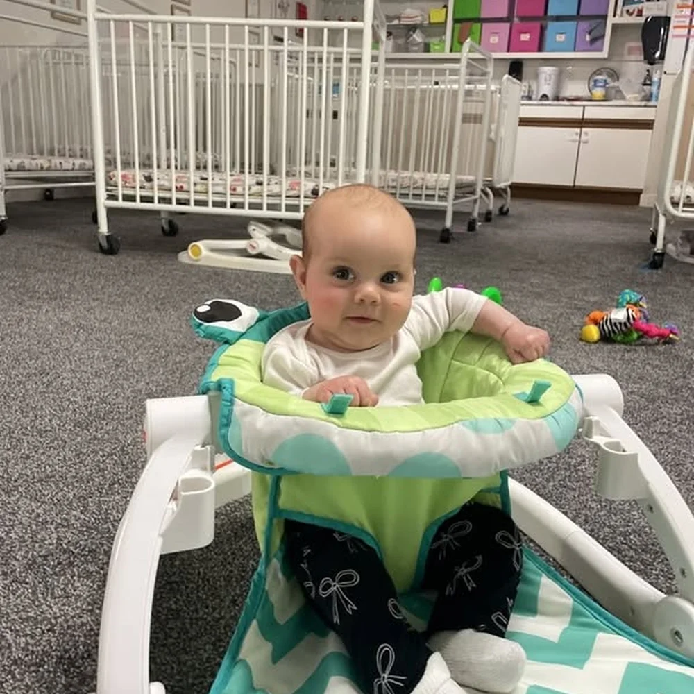 Smiling baby sitting supported in a green floor seat in the Owlets infant room