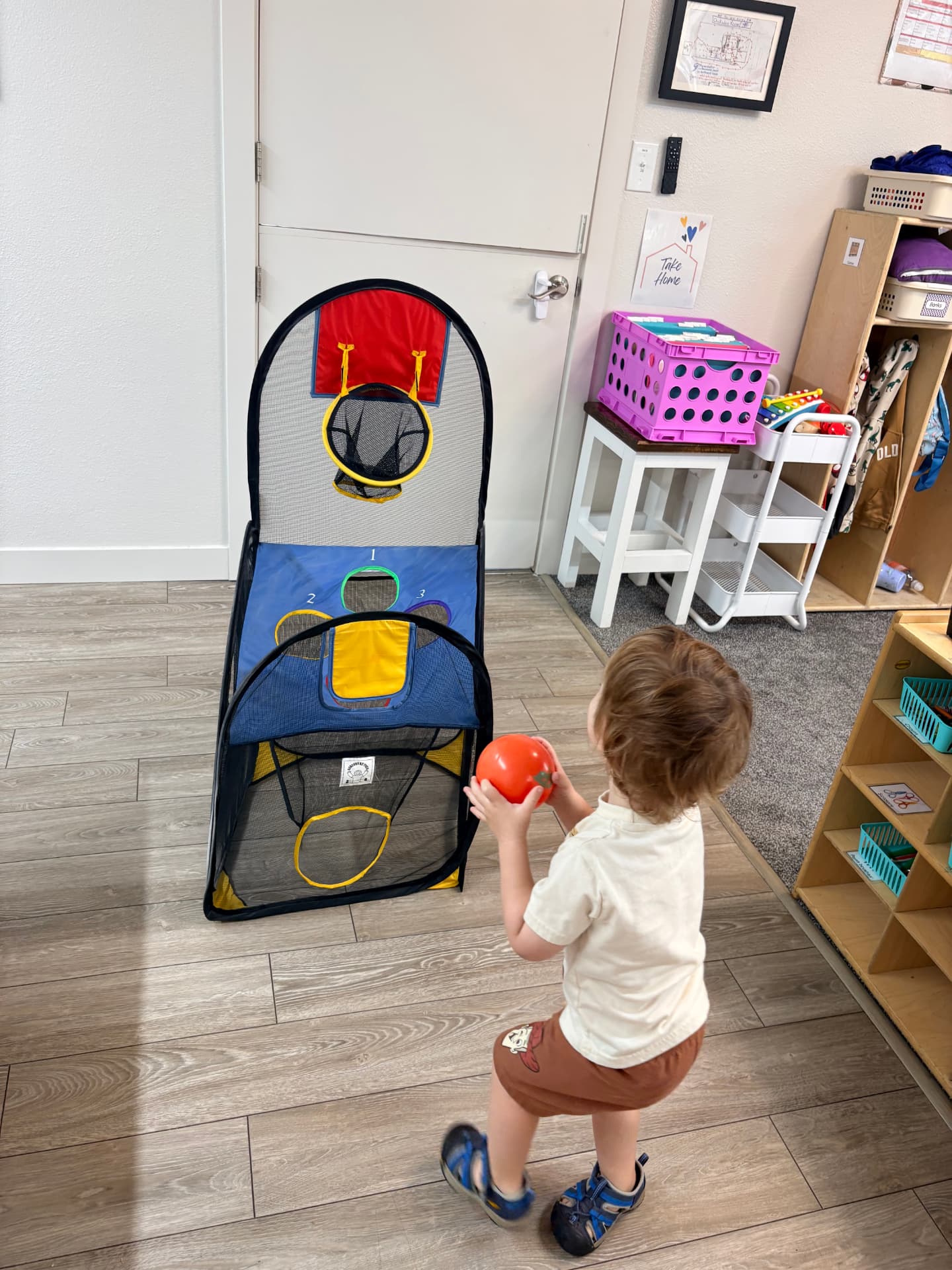 Toddler in a cream shirt carrying an orange ball toward the indoor basketball and bean bag toss game