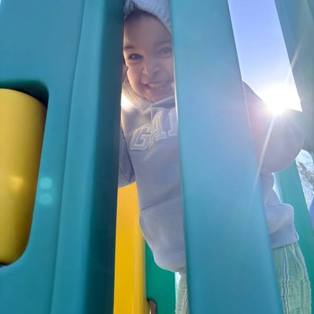 A bundled up toddler smiling through the bars of a playground climber on a sunny winter day