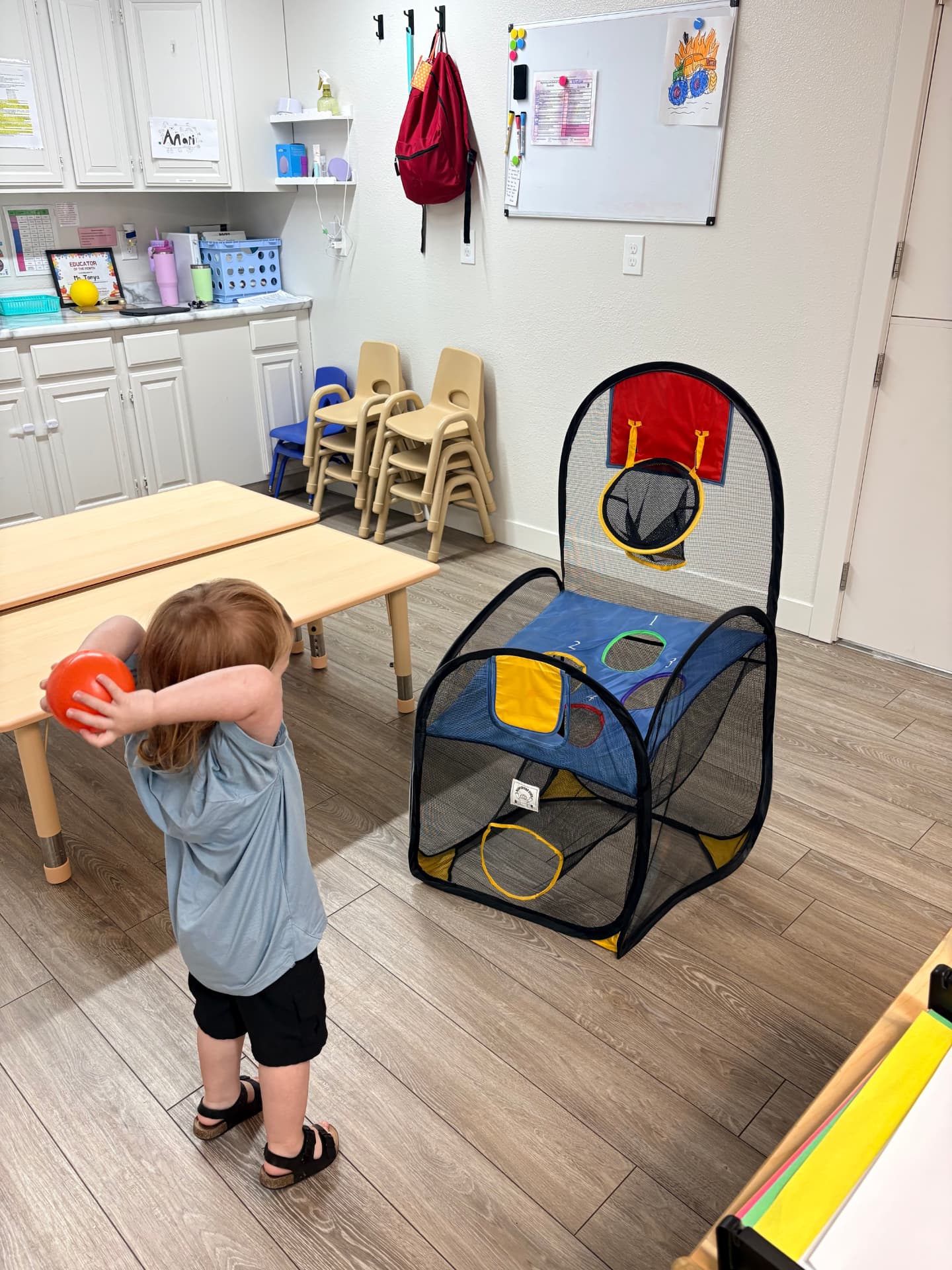 Toddler girl winding up to toss an orange ball at an indoor pop-up basketball hoop in the Chickadees classroom