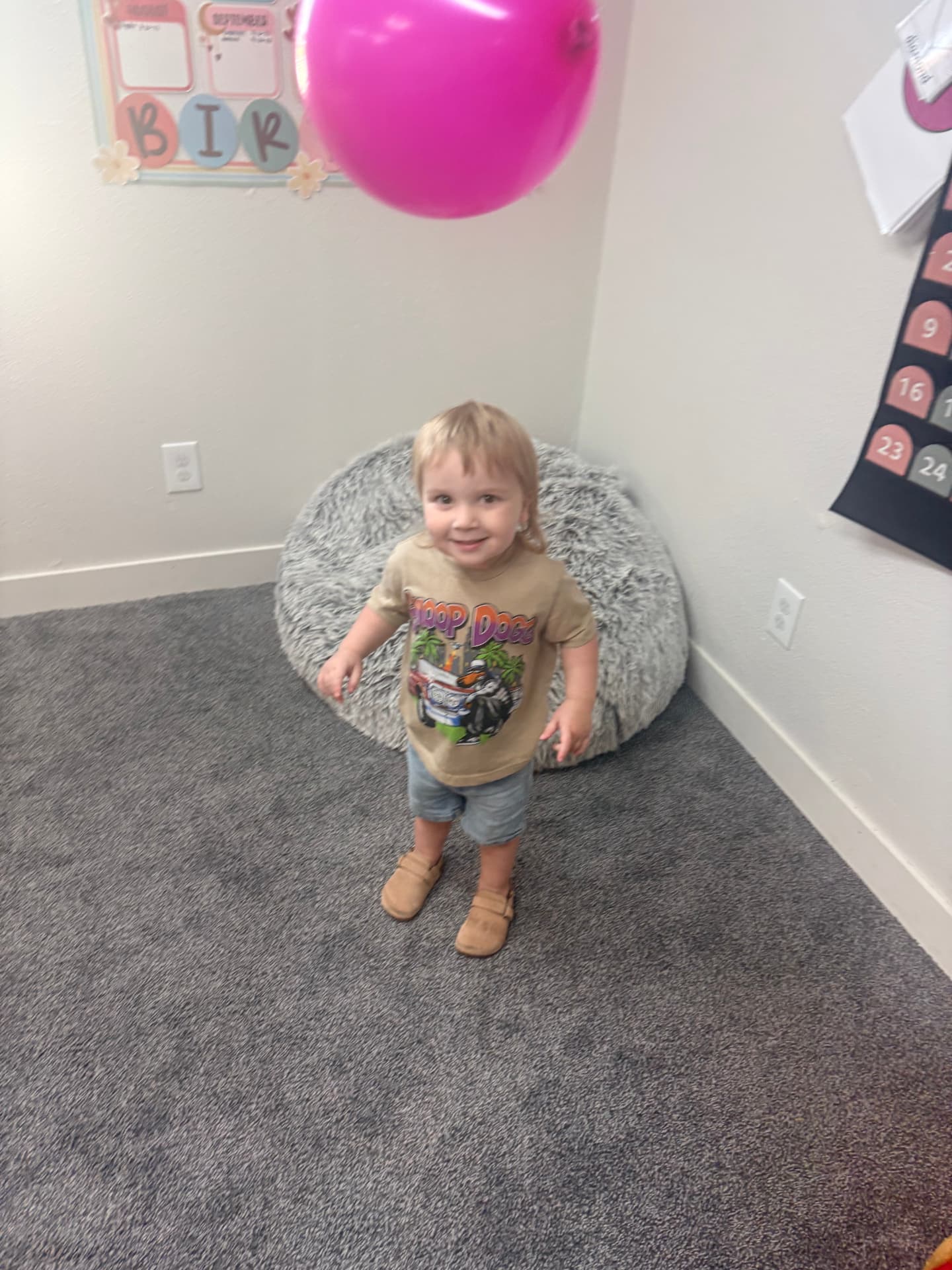 Smiling toddler standing under a floating pink balloon in the Chickadees cozy corner at The Nest