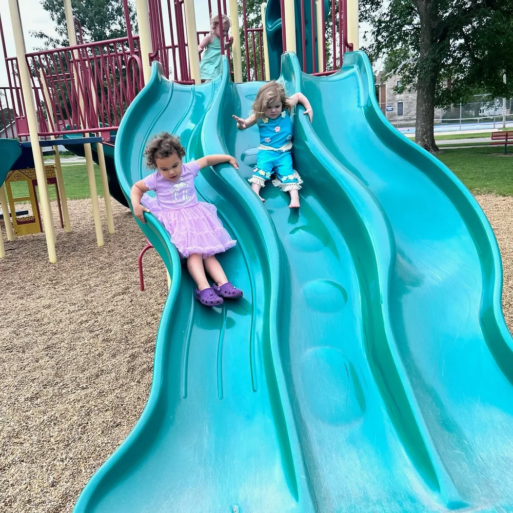 Two young girls sliding down a turquoise playground slide at a park near The Nest in Earlham, Iowa
