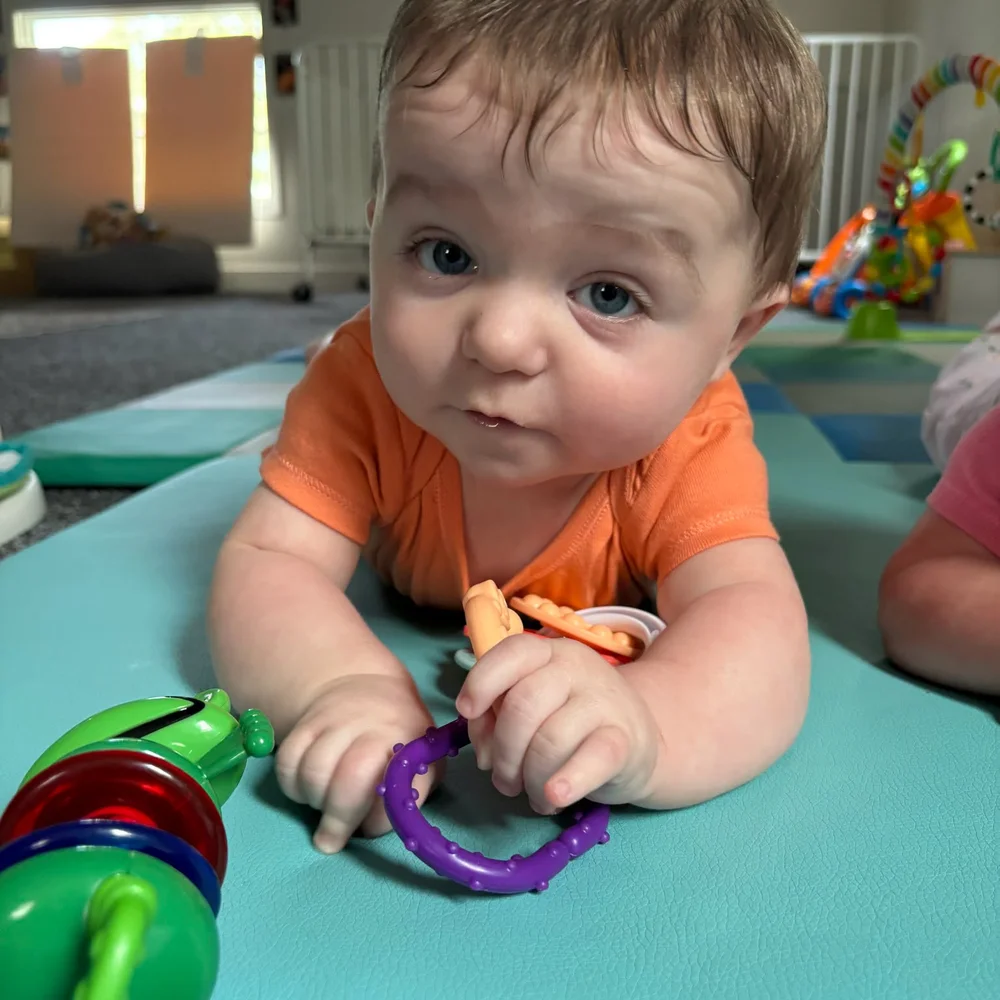 Infant in an orange shirt during tummy time on a soft teal mat, holding a purple teething ring
