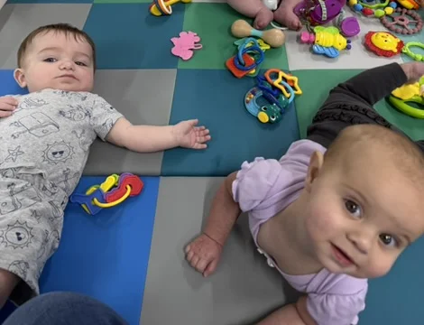 Two infants on a colorful checkered foam play mat reaching toward bright toys at The Nest Owlets room