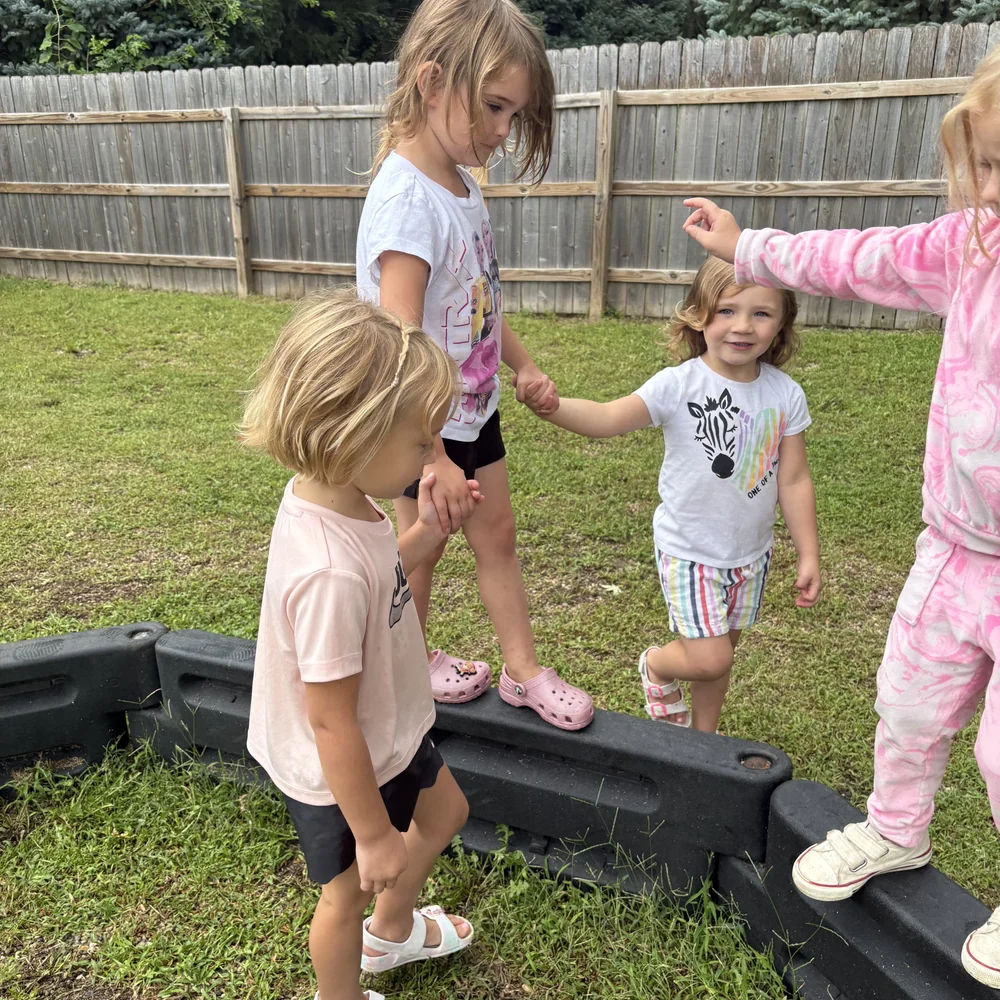 Preschoolers holding hands and balancing along an outdoor sandbox edge in the fenced playground at The Nest