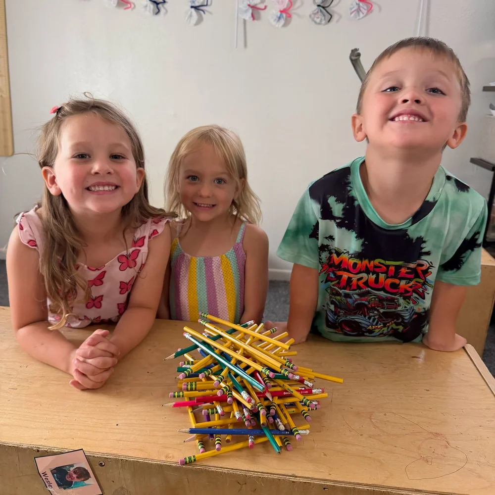 Three smiling children at a table with colorful pencils in a classroom at The Nest Child Care and Early Learning Center