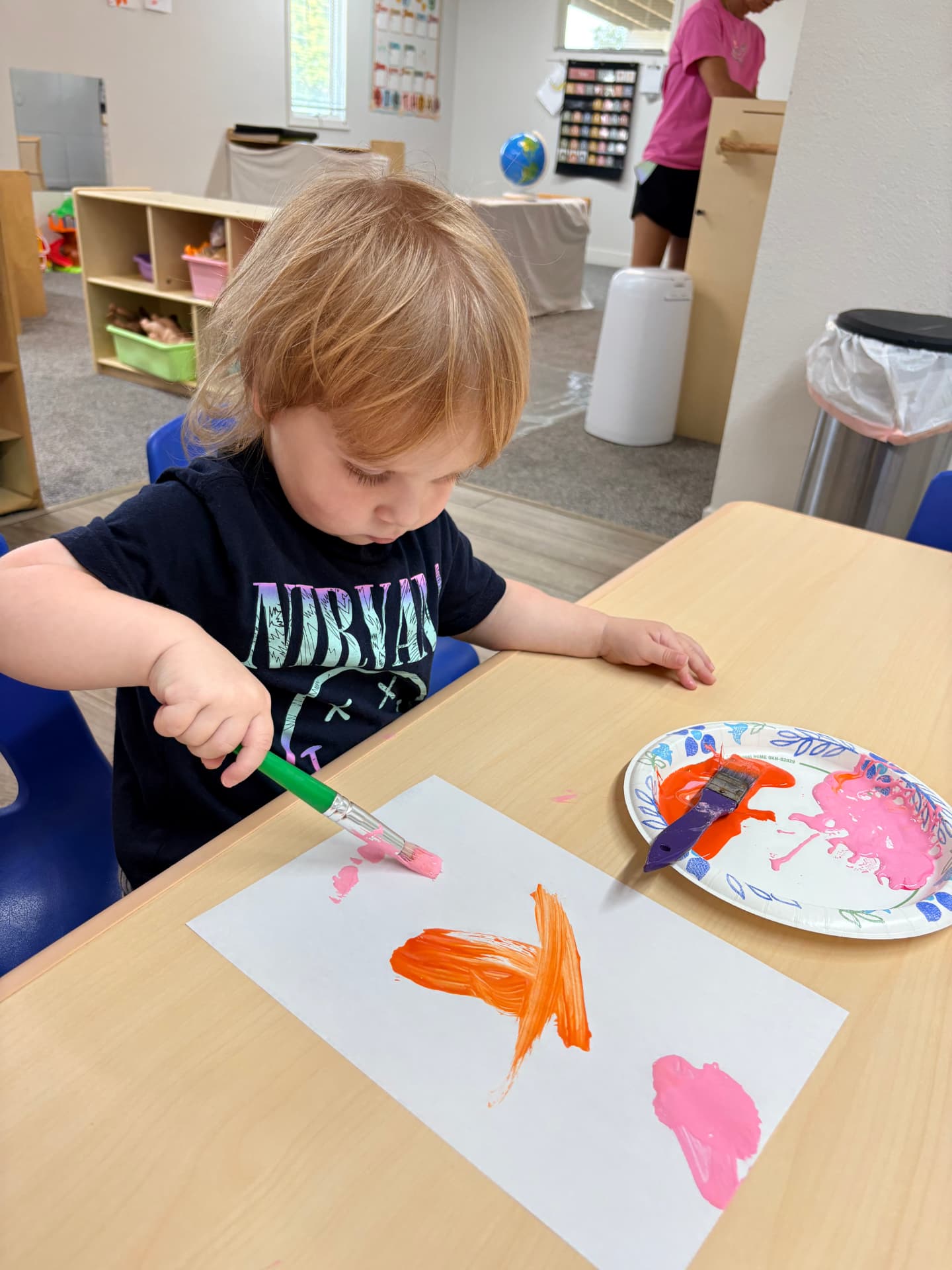 Toddler boy in a black t-shirt painting orange and pink brushstrokes with a wide brush at a classroom table