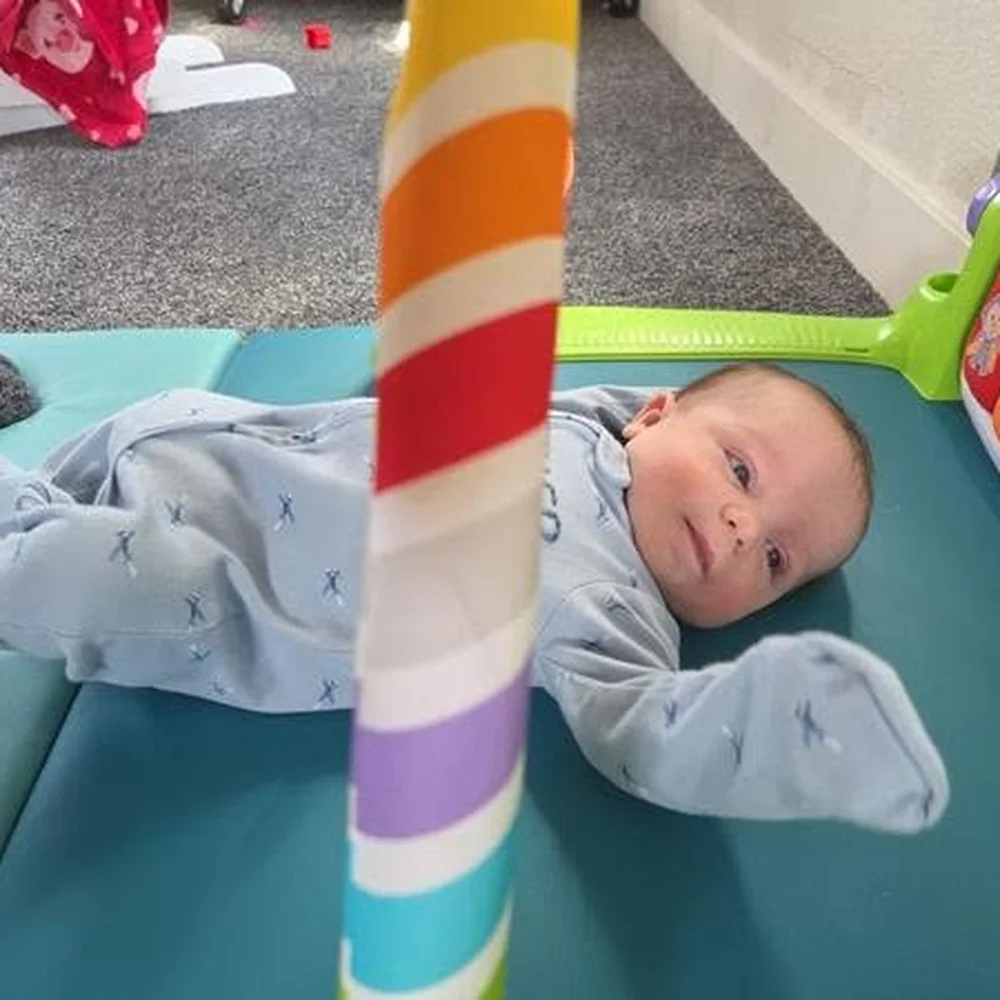 Baby in a blue sleeper resting on a padded play mat under a colorful striped activity gym
