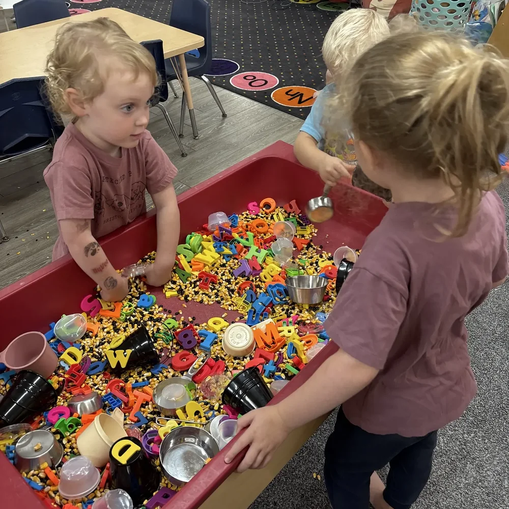 Preschoolers scooping magnetic letters and corn kernels at the red sensory table at The Nest