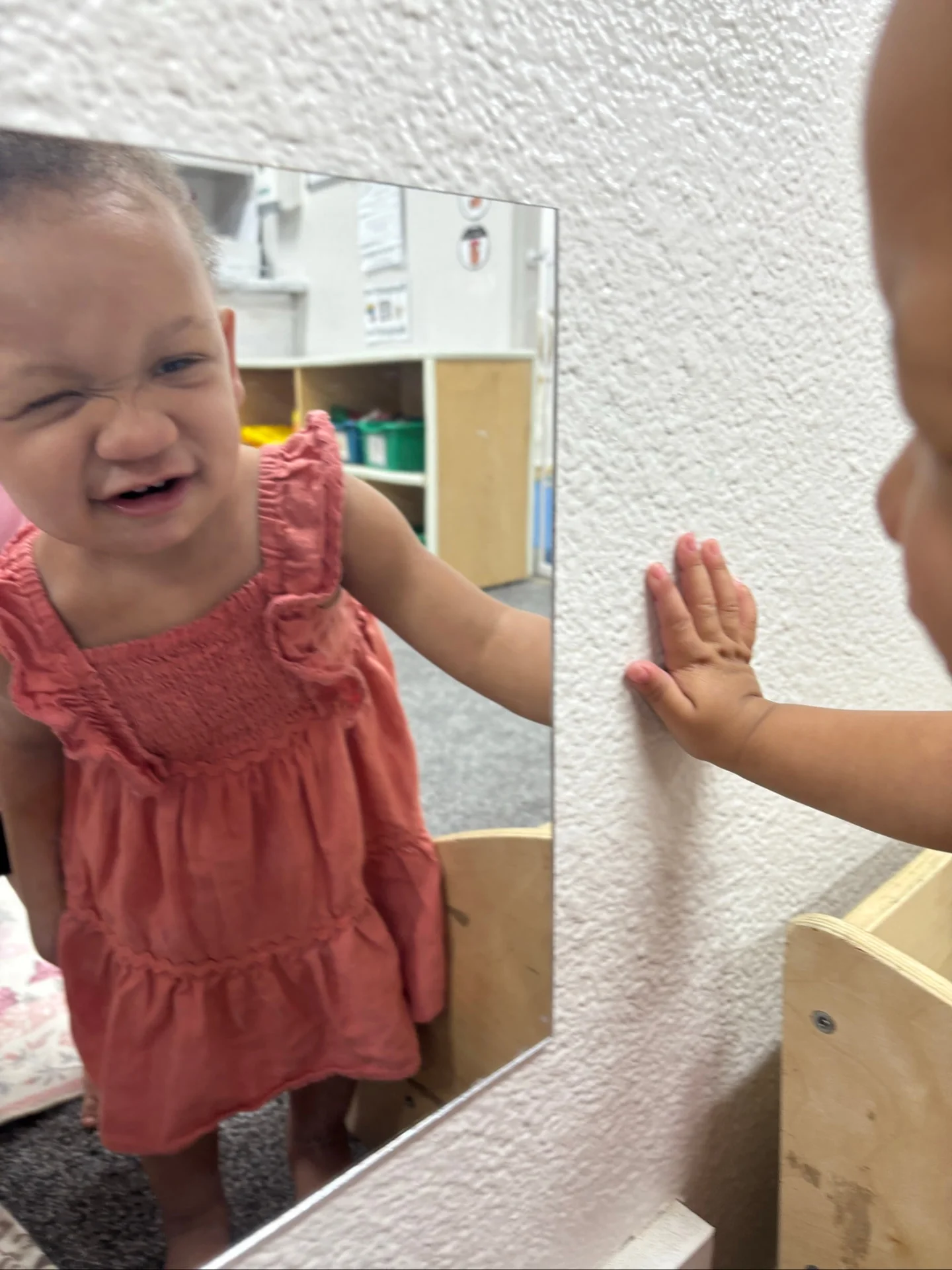 Smiling toddler in a coral dress playing at a mirror in the Flamingos room