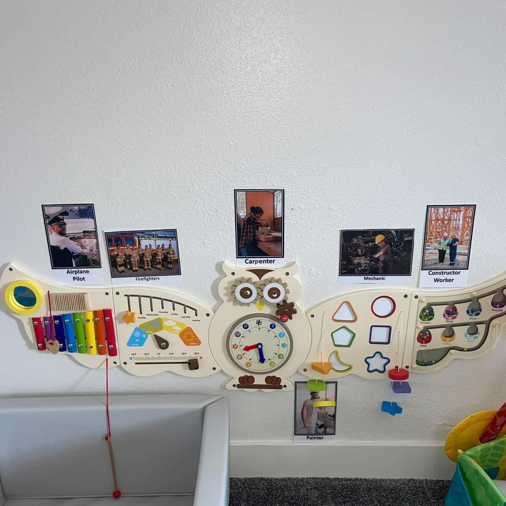 Wooden owl shaped sensory activity board with xylophone, clock, shapes, and career photo cards on the infant room wall