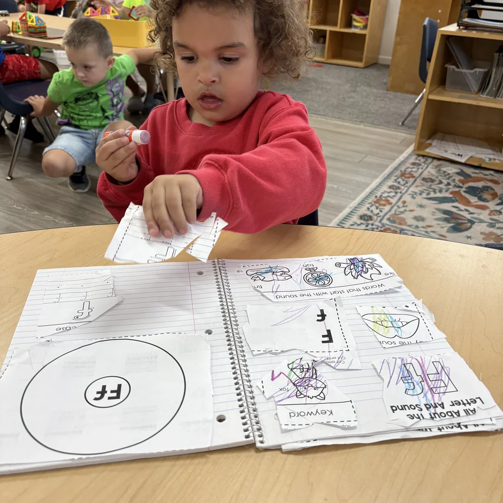 Preschooler gluing letter F cutouts into a phonics notebook during a Heggerty literacy lesson at The Nest