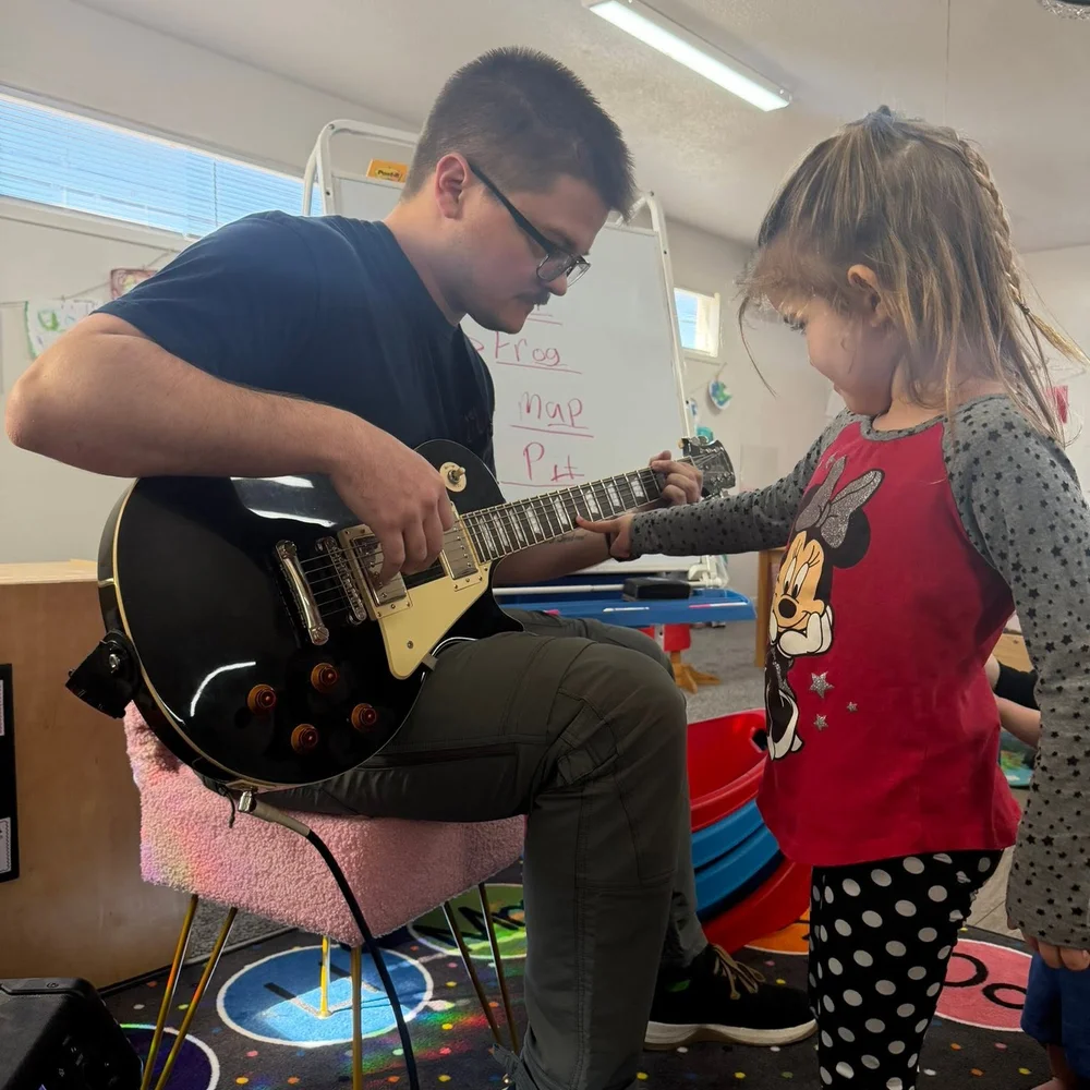 A music visitor playing electric guitar while a preschool girl strums the strings at The Nest