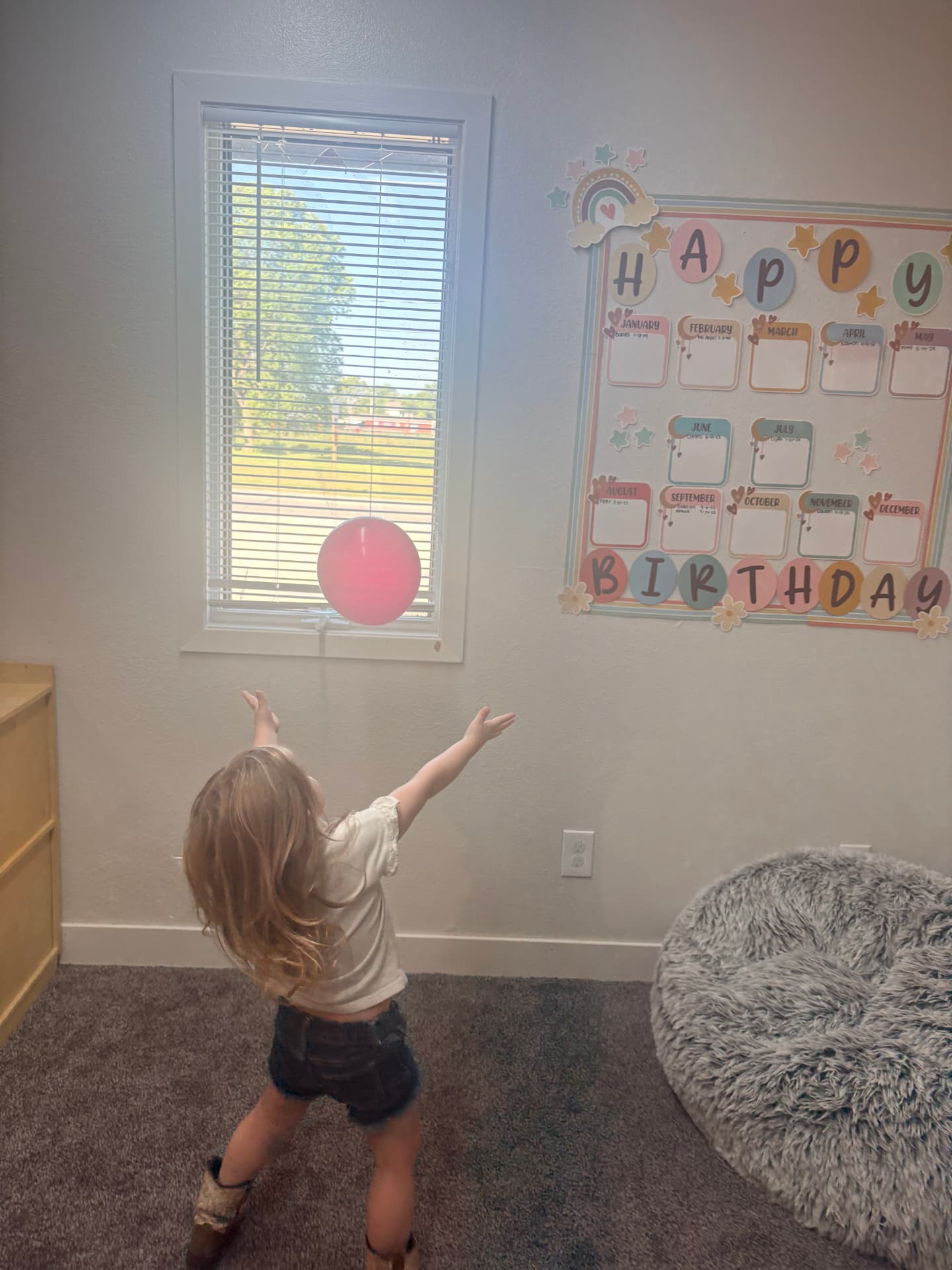 Toddler girl tossing a pink balloon into the air beside the classroom birthday board at The Nest