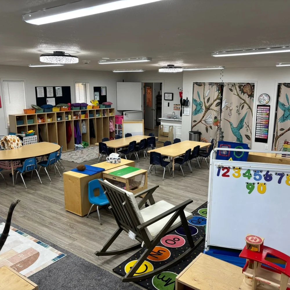 Full preschool classroom with tables, cubbies, hummingbird curtains, and a number board