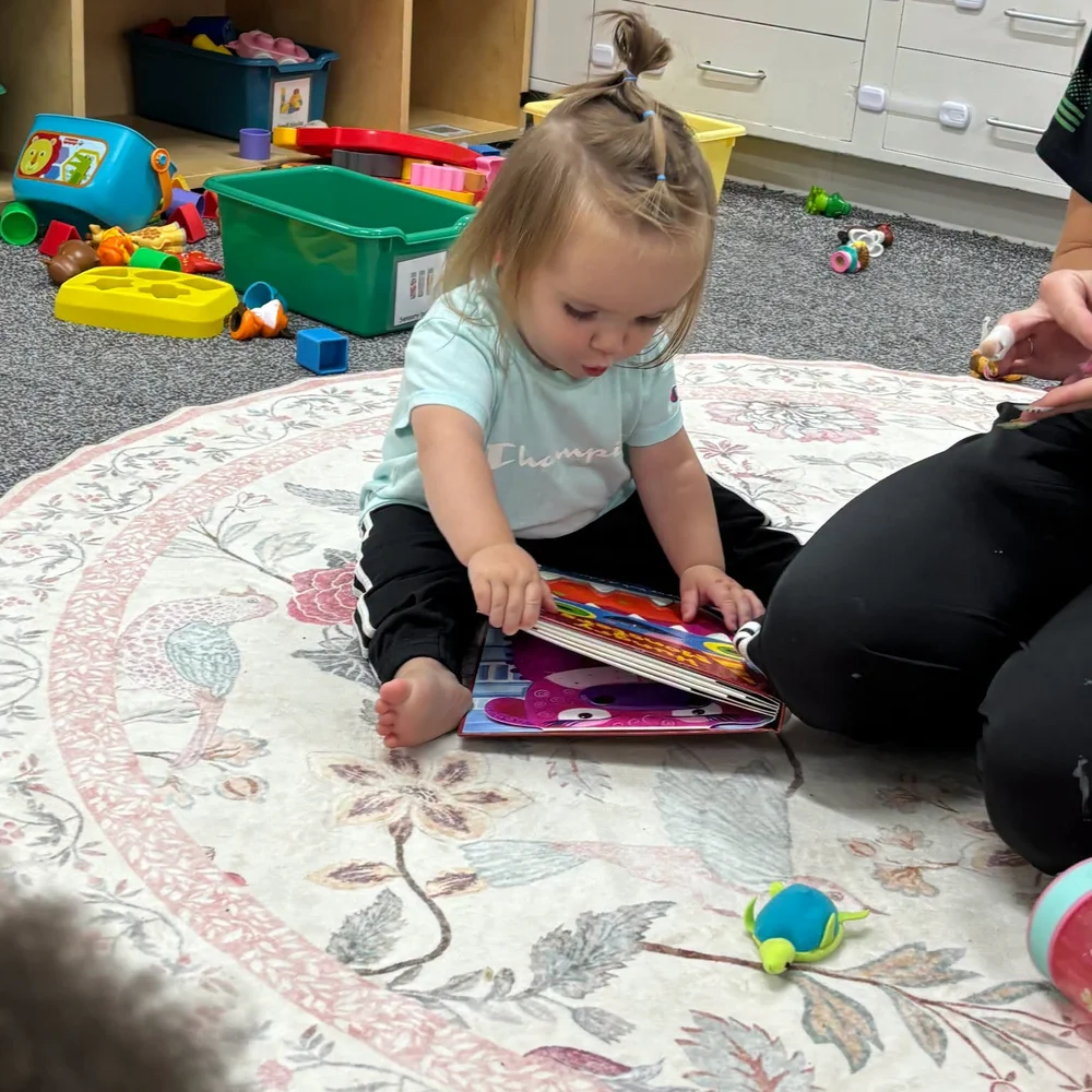 Toddler sitting on a floral rug turning the pages of a colorful board book