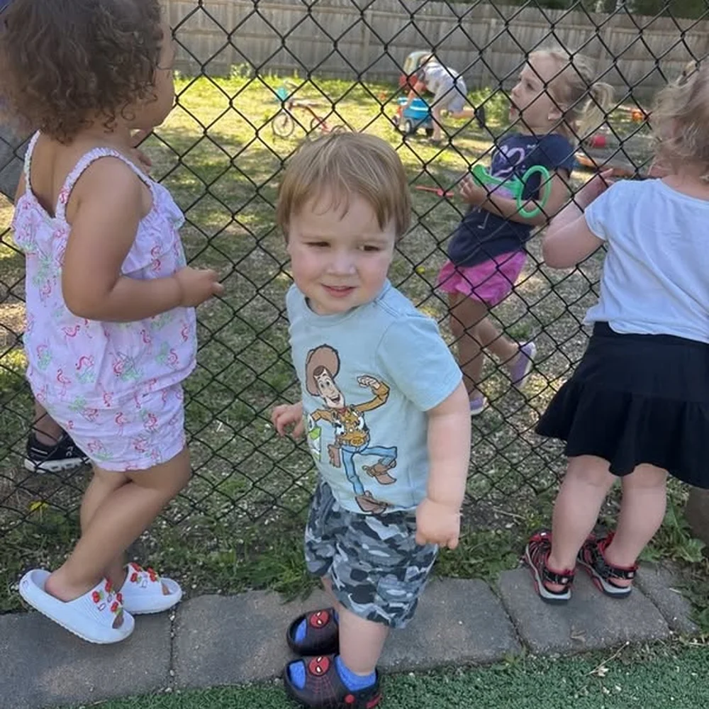 Toddlers playing outside along the fence of the grassy play yard at The Nest