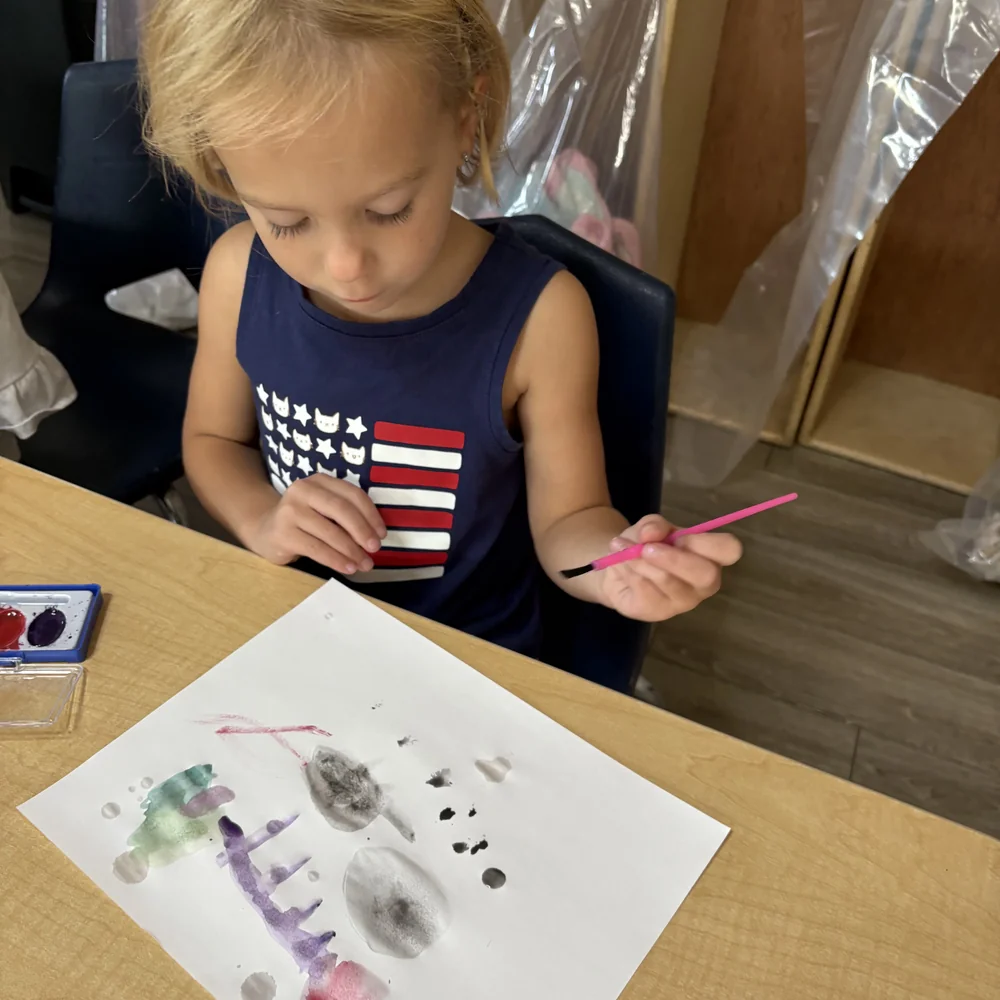 Preschool girl concentrating on a watercolor painting at a classroom table at The Nest