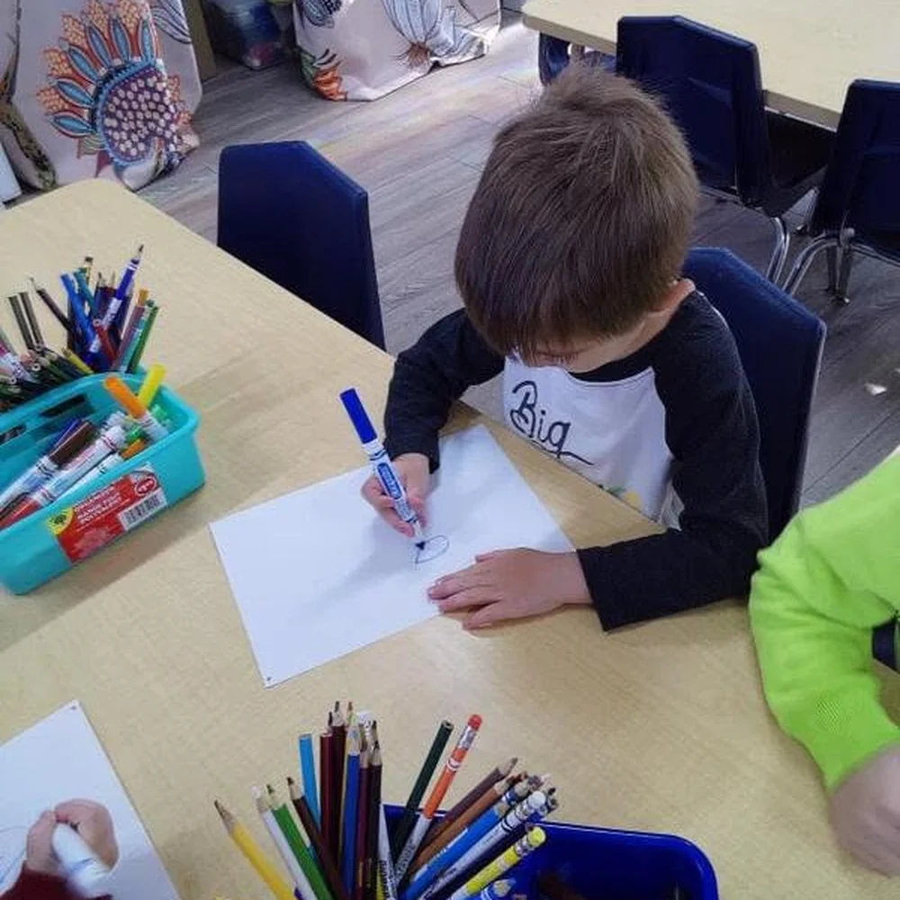 Preschool boy drawing with a blue marker at a classroom table full of pencils and markers at The Nest
