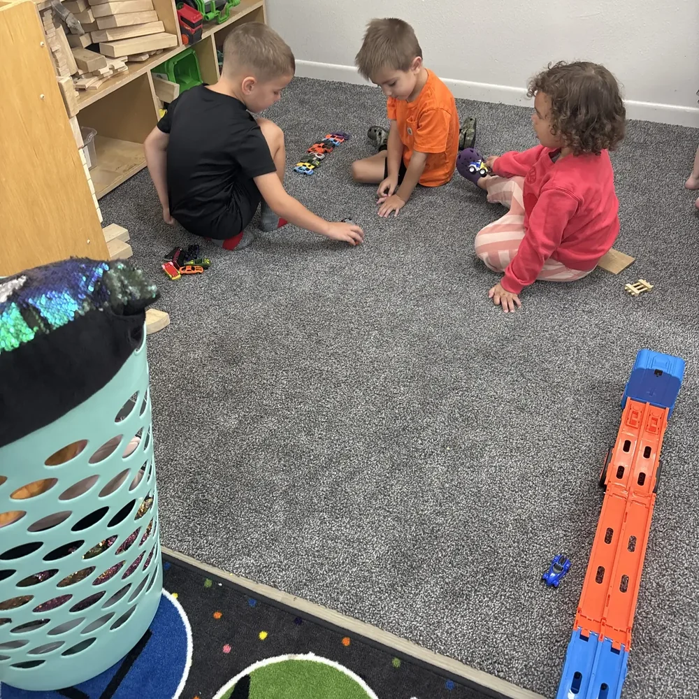 Three preschoolers sitting on the classroom carpet playing with toy cars and a race track at The Nest