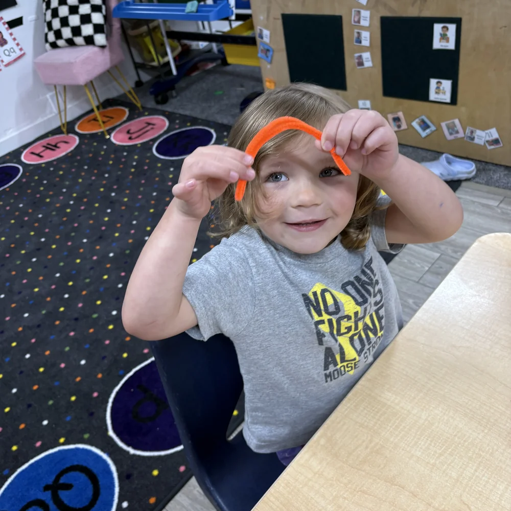 Preschool boy smiling while bending an orange pipe cleaner during a fine motor activity at The Nest