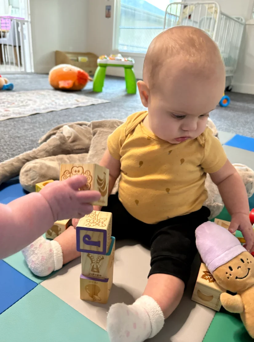Baby in a yellow shirt stacking wooden alphabet blocks with a caregiver's hand nearby in the Owlets infant room
