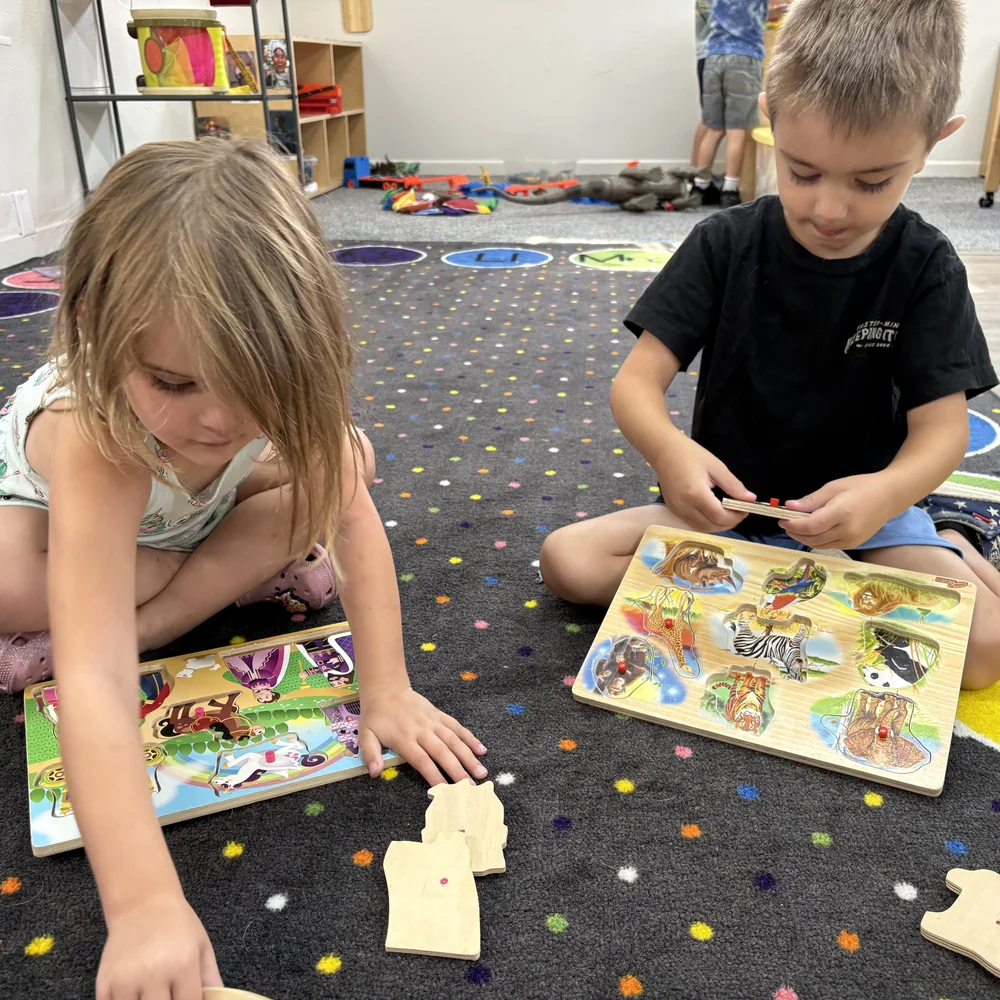 Two preschoolers working on wooden animal puzzles on the alphabet rug in the Hummingbirds classroom