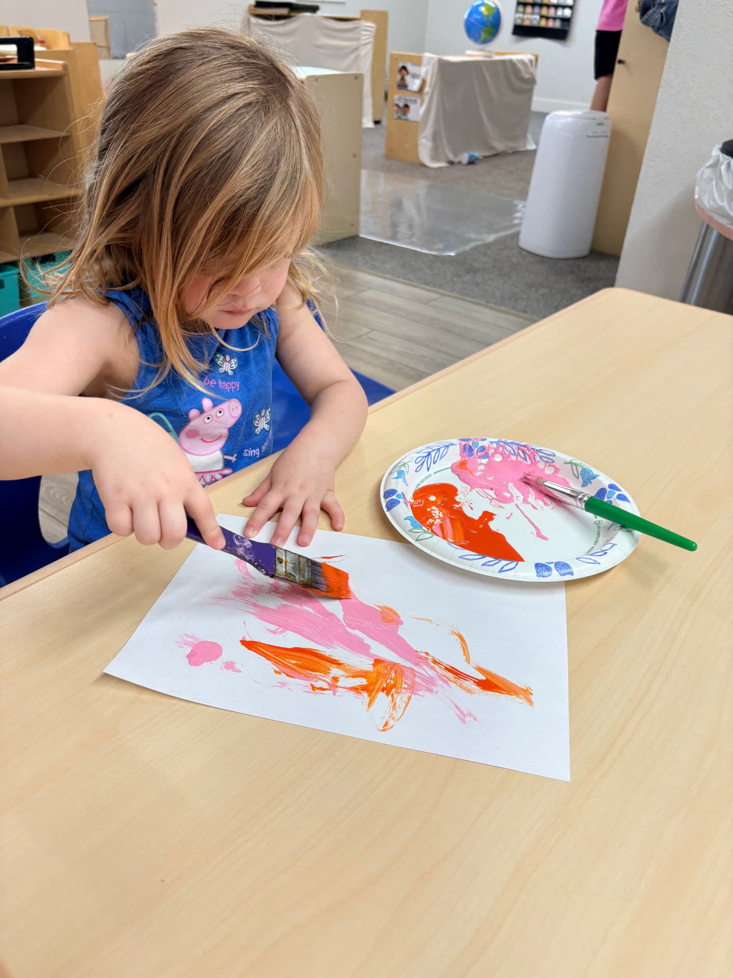 Toddler girl in a blue shirt spreading pink and orange paint across her paper during art time