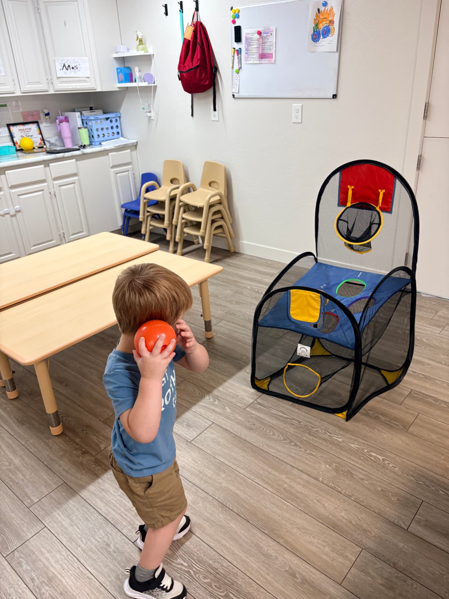 Toddler boy holding an orange ball and aiming at the pop-up basketball hoop across the classroom