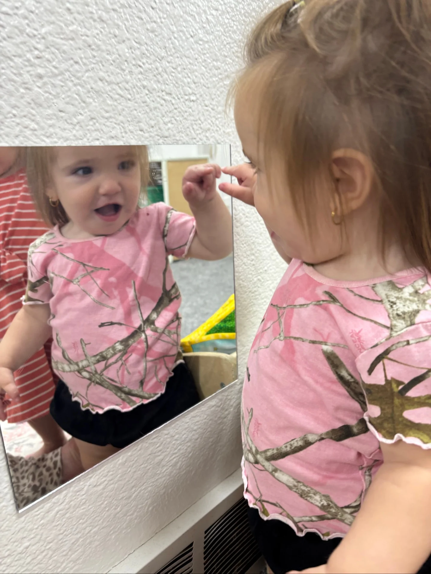 Toddler in a pink shirt discovering her reflection in a mirror at The Nest