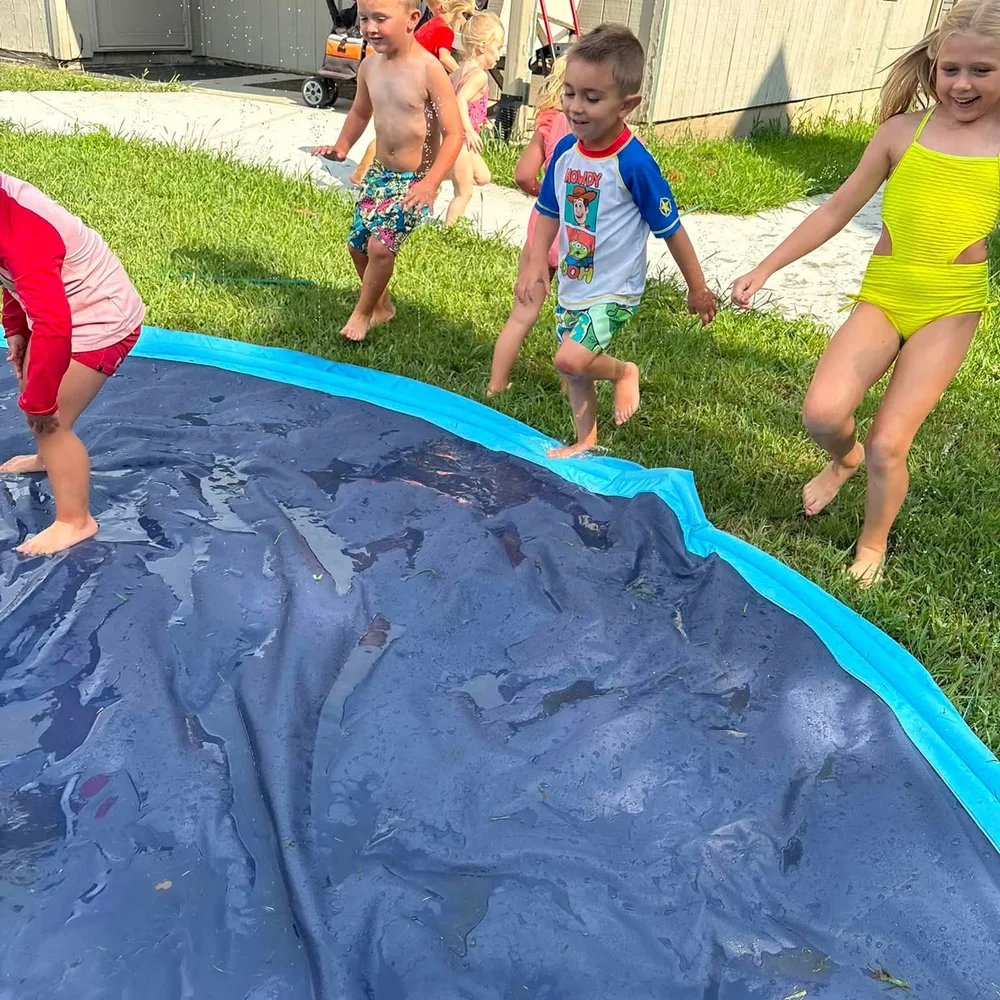 Children splashing and running across a water slip mat during summer outdoor play at The Nest
